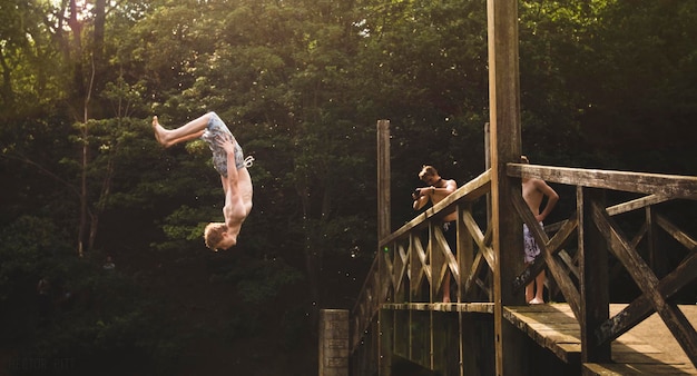 friend photographing man diving from wooden bridge against trees in forest