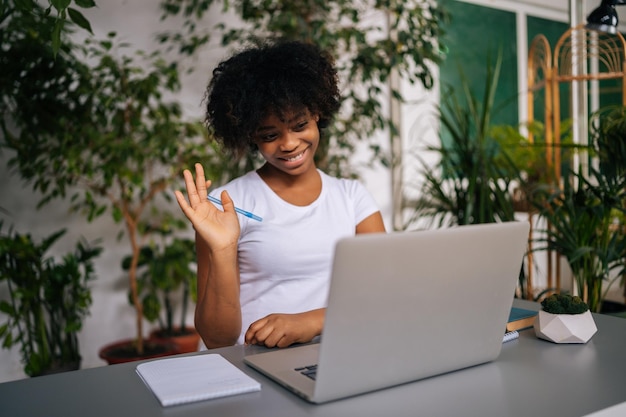 친절한 아프리카계 미국인 여학생이 가벼운 집에서 교사와 화상 회의에서 말하는 노트북의 웹캠에서 손을 흔들며 인사합니다.