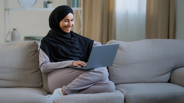 Friendly arabian muslim woman girl in hijab sit on cozy sofa talk video call with friend greeting at