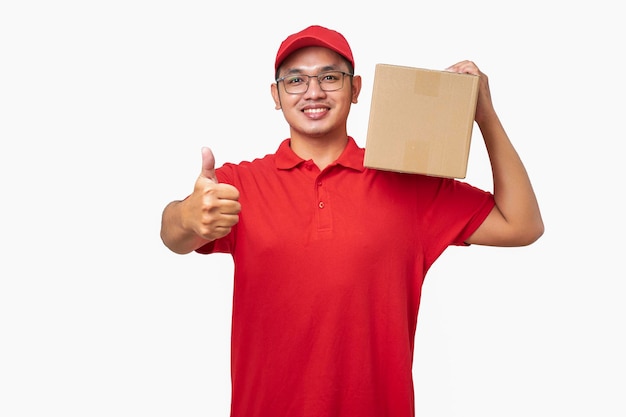 Friendly asian courier delivery man wearing red shirt and cap showing thumbup holding box package on shoulder