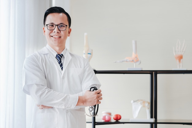 Friendly asian doctor olding stethoscope in hand in his office