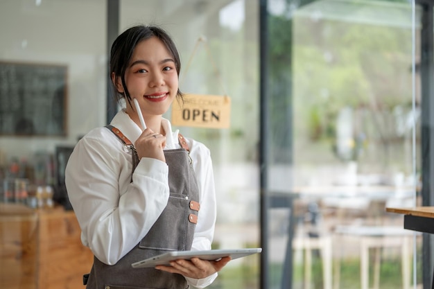 A friendly Asian female cafe worker or waitress stands in her cafe with a tablet in her hand