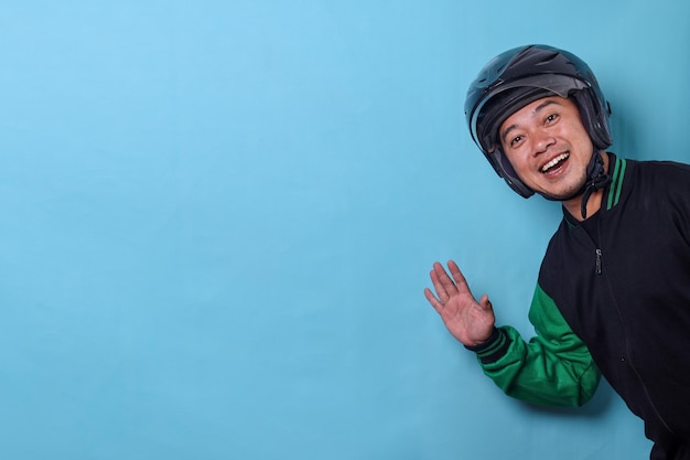 Friendly Asian online taxi driver wear helmet peeking and waving hand isolated on blue background wi