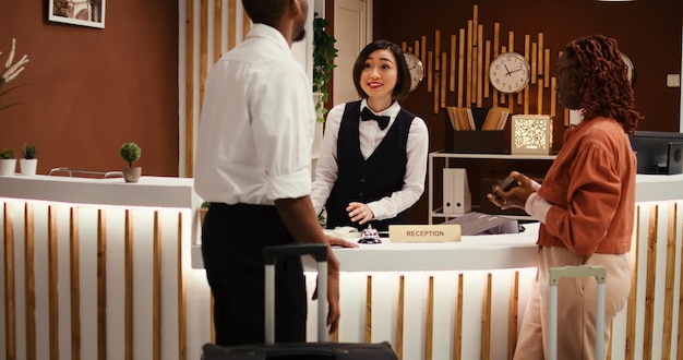 Friendly asian receptionist assisting guests with booking reservation, inviting them to wait in stylish cozy hotel lounge. Happy guests helped by bellboy with their luggage