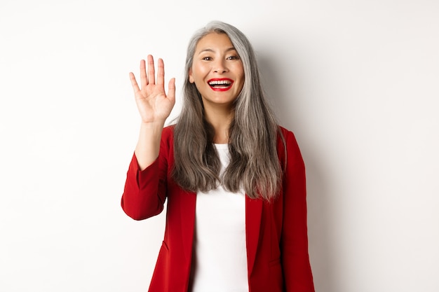 Friendly asian senior lady in elegant blazer saying hi, waving hand and greeting you with happy smile, standing over white background.