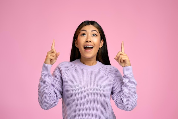 Friendly asian woman points fingers up at empty space isolated on pink background