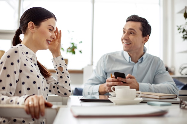 Friendly atmosphere. Handsome cheerful dark-haired man smiling and holding his phone and his female colleague sitting next to him and they talking