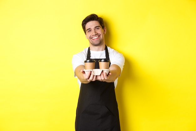 Barista amichevole in grembiule nero che dà ordine da asporto, con in mano due tazze di caffè e sorridente, in piedi sopra il muro giallo