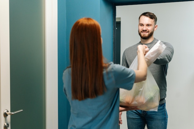 Friendly bearded courier male delivering groceries bag to unrecognizable female housewife customer on doorway at home