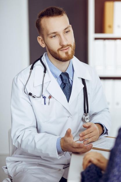 Friendly bearded doctor and patient woman discussing current health examination while sitting in sunny clinic. Medicine concept