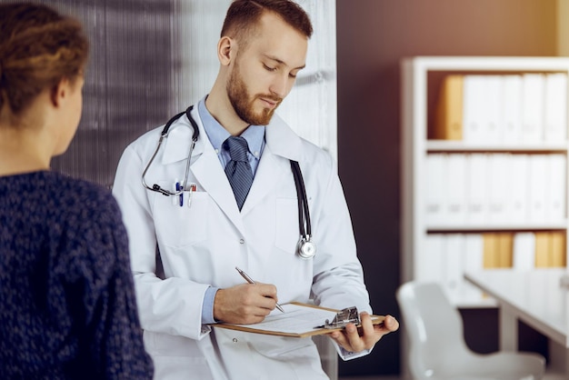 Friendly bearded doctor and patient woman discussing current health examination while sitting in sunny clinic. Medicine concept.