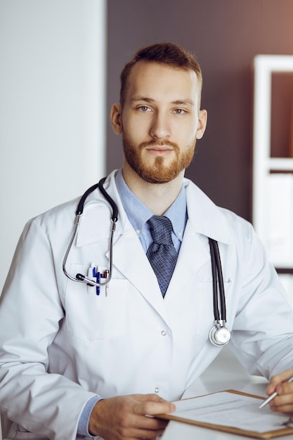 Friendly bearded doctor sitting and writing at clipboard in sunny clinic. Medicine concept.