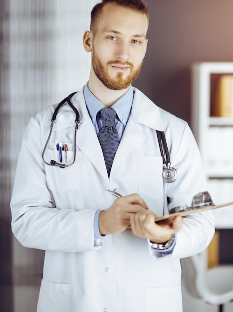 Friendly bearded doctor standing and writing with clipboard in sunny clinic at his working place. Medicine concept