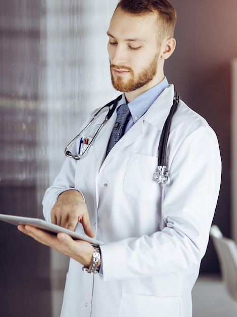 Friendly bearded doctor using tablet computer in sunny clinic at his working place. Medicine concept.