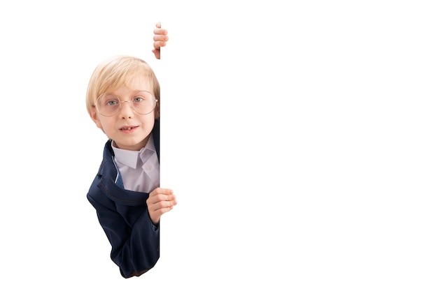 Friendly blond boy in glasses peeking from behind white backdrop Schoolboy advertising your text Copy space