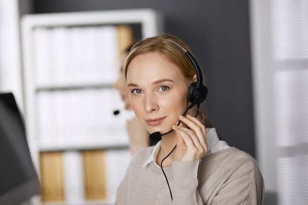 Friendly businesswoman talking by headset while sitting in office. Call center and diverse people group in business.