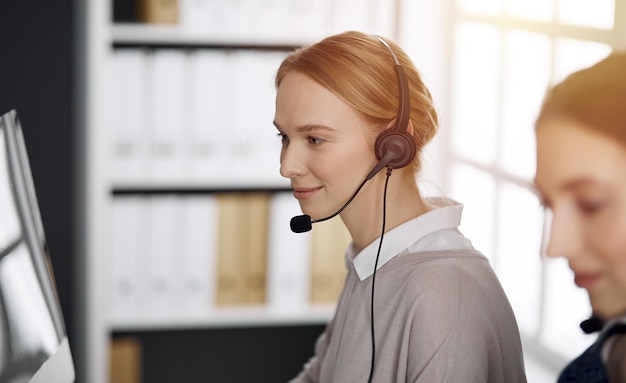 Friendly caucasian businesswoman talking by headset in sunny office. Call center and diverse people group in business.