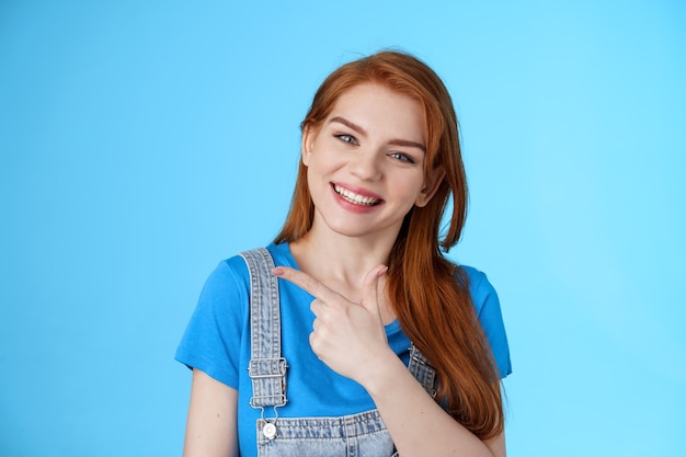 Friendly cheerful caucasian red woman, tilt head smiling pleasantly, suggest good copy space, pointing left introduce promo, give advice, indicating awesome link, pointing blue background