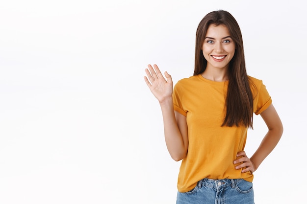 Amichevole allegra, donna sorridente felice che ti saluta con la mano alzata. ragazza attraente che saluta amico, saluta o ciao, benvenuto ospite, in piedi sfondo bianco gioioso, esprimi positività e gioia