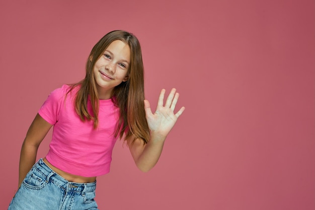 Photo friendly child girl blogger waving hand in greeting gesture smiling welcoming subscribes on pink