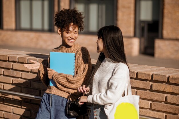 Friendly conversation. Two smiling gesturing girls with bag and notebook having fun talking outdoors standing in office area on sunny day