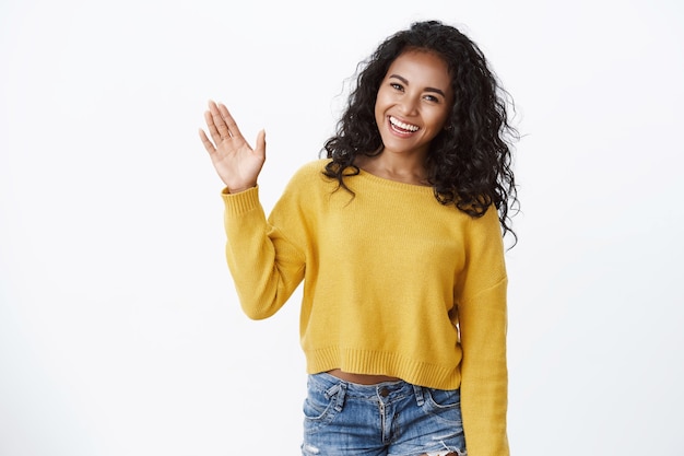 Foto amichevole ragazza dai capelli ricci carina in maglione giallo alza il palmo e saluta, saluta, saluta gli ospiti inclinando la testa adorabile sorridente, fa un gesto di addio, in piedi muro bianco