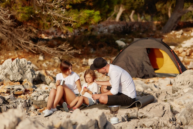 Photo friendly family, sitting near tent on stone seacoast at daytime.