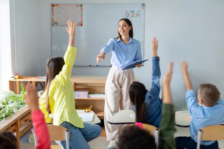 Photo friendly female teacher in classroom teaches elementary school students kids raising hands to