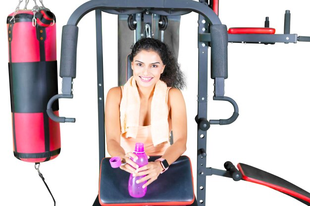 Дружная фитнес-молодая индийская женщина с бутылкой воды и улыбается с тренажерным оборудованием, изолированным на белом