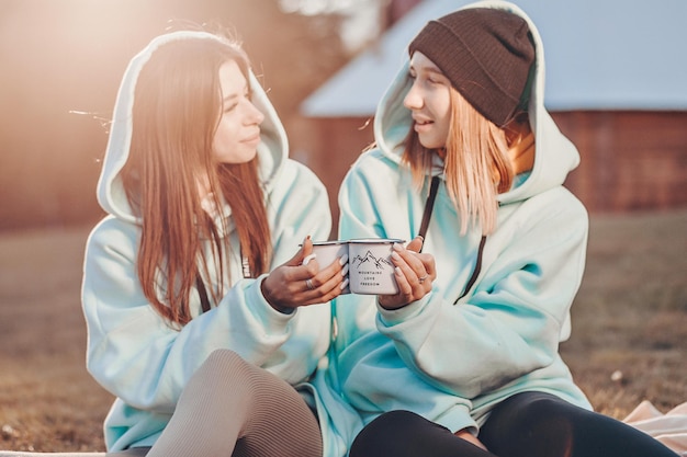 Friendly gatherings in a warm atmosphere. Girls in identical blue hoodies sit on a blanket with mugs of tea and communicate with each other. Next to a wooden house.
