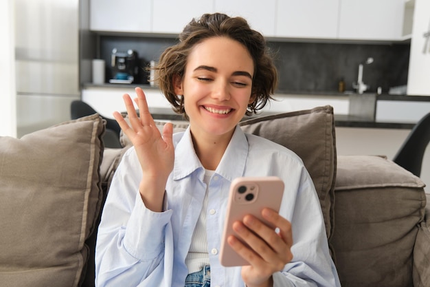 Friendly girl talks on mobile phone video chats with friend waves hand at smartphone camera sits on