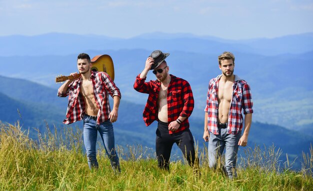 晴れた日にギターハイキングをするフレンドリーな男たち一緒に自由を楽しむ山の頂上を一緒に歩く市松模様のシャツを着た若者のグループ観光客のハイキングのコンセプト友達とのハイキング