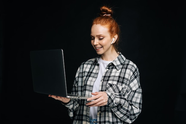 Friendly happy young business woman or student holding laptop computer and looking at display on isolated black background Pretty lady model with red hair emotionally showing facial expressions