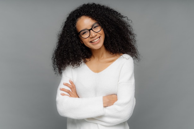 Friendly looking African American woman tilts head, has dark crisp bushy hair, keeps arms folded over chest