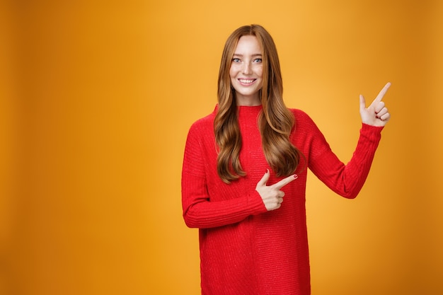 Приветливая, веселая и энергичная рыжая женщина в красном свитере указывает на верхний правый угол, продвигая рекламу с широкой восторженной и нежной улыбкой, позирующей на оранжевом фоне