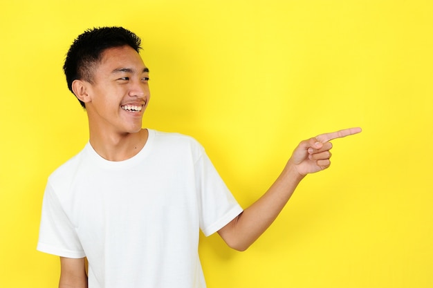 Friendly-looking lively pleasant Asian male with white t-shirt smiling delighted look at copy space joyful. Self-assured pointing, on yellow background