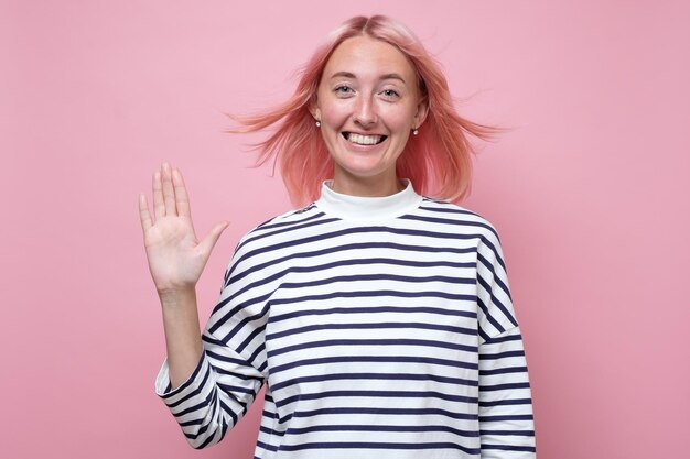 Friendly looking young woman with pink dyed hair smiling happily saying Hello