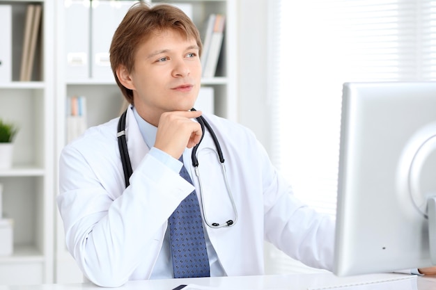 Friendly male doctor is sitting at the table and working in the hospital office Ready to examine and help patients High level and quality medical service concept