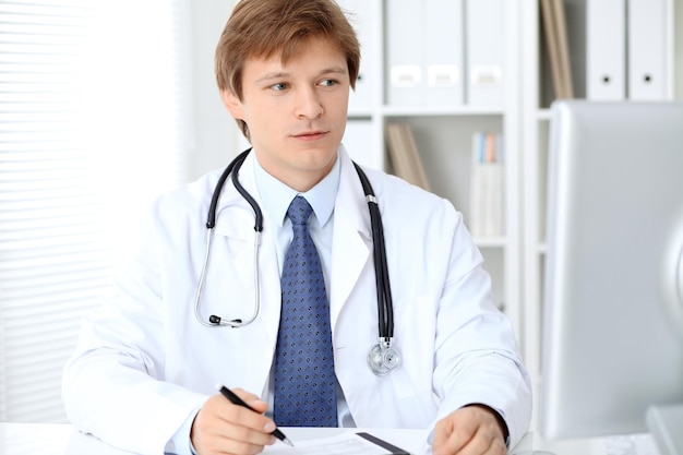 Friendly male doctor is sitting at the table and working in the hospital office. Ready to examine and help patients. High level and quality medical service concept.