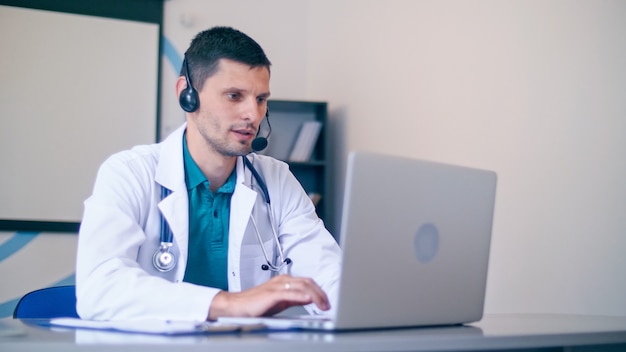 Friendly Male Doctor in White Medical Coat With Headphones Making Conference Call on Laptop.Remote Consulting Patient Online From Healthcare Hospital. Telemedicine.