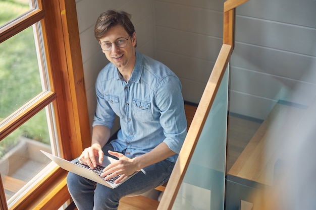 дружелюбный мужчина сидит на деревянной лестнице и работает за серым ноутбуком