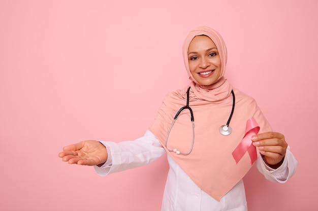 ピンクのヒジャーブと首の周りの聴診器で、ピンクのリボンを保持し、色付きの背景に手で示しているフレンドリーな中東民族のイスラム教徒の女性。乳がん啓発キャンペーン、コピースペース