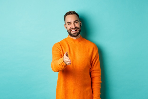 Friendly nice guy extend hand for handshake, greeting you and smiling, saying hello, standing over light blue background.