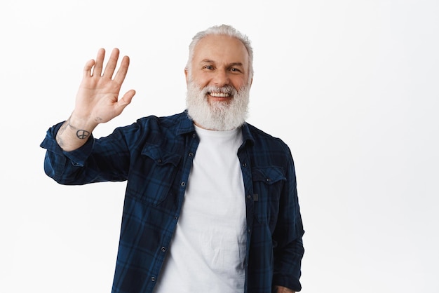 Amichevole vecchio agitando la mano per dire ciao ragazzo anziano sorridente con tatuaggi onde come saluto facendo ciao gesto in piedi felice su sfondo bianco