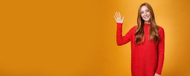 Friendly and optimistic good-looking ginger girl in red sweater raising palm waving at camera person