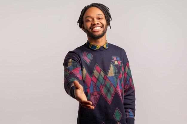 Photo friendly positive african man with dreadlocks in trendy sweatshirt holds out his hand with toothy smile introducing himself nice to meet you indoor studio shot isolated on gray background