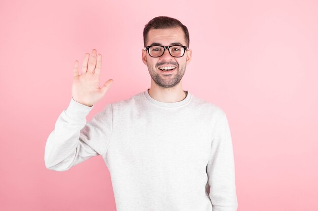 Friendly positive smiling young man with stubble and dark hair in a trendy sweatshirt waving hand while greeting friends while having fun indoors