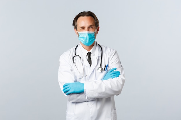 Friendly professional doctor in medical mask and gloves, encourage patient relax, all good, winking and cross arms chest