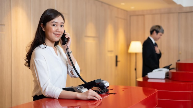 Friendly receptionist woman working and call talking to customer by telephone at desk in hotel lobby