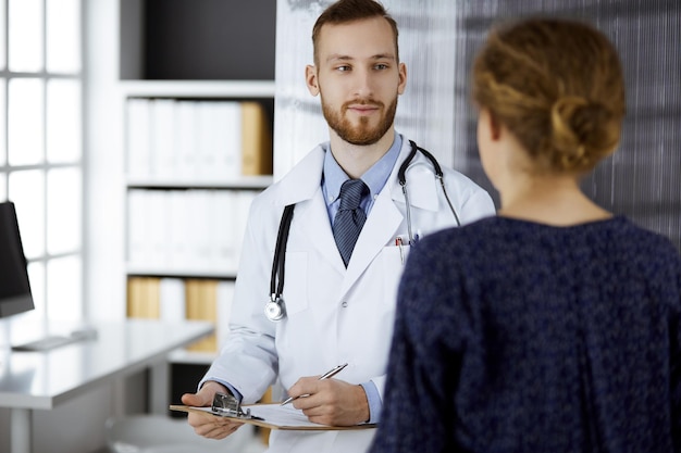 Friendly red-bearded doctor and patient woman discussing current health examination while sitting in clinic. Perfect medical service and medicine concept.